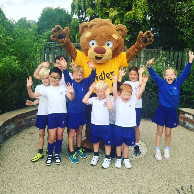 Small school children with their hands in the air with Sidley the bear